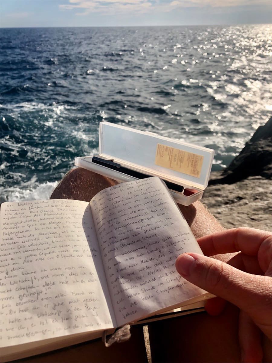 Eine Hand hält ein offenes Notizbuch am Meer.