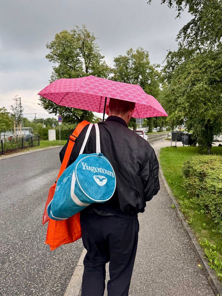 Ein von hinten fotografierter Mann hält einen Regenschirm und trägt diverse Taschen.