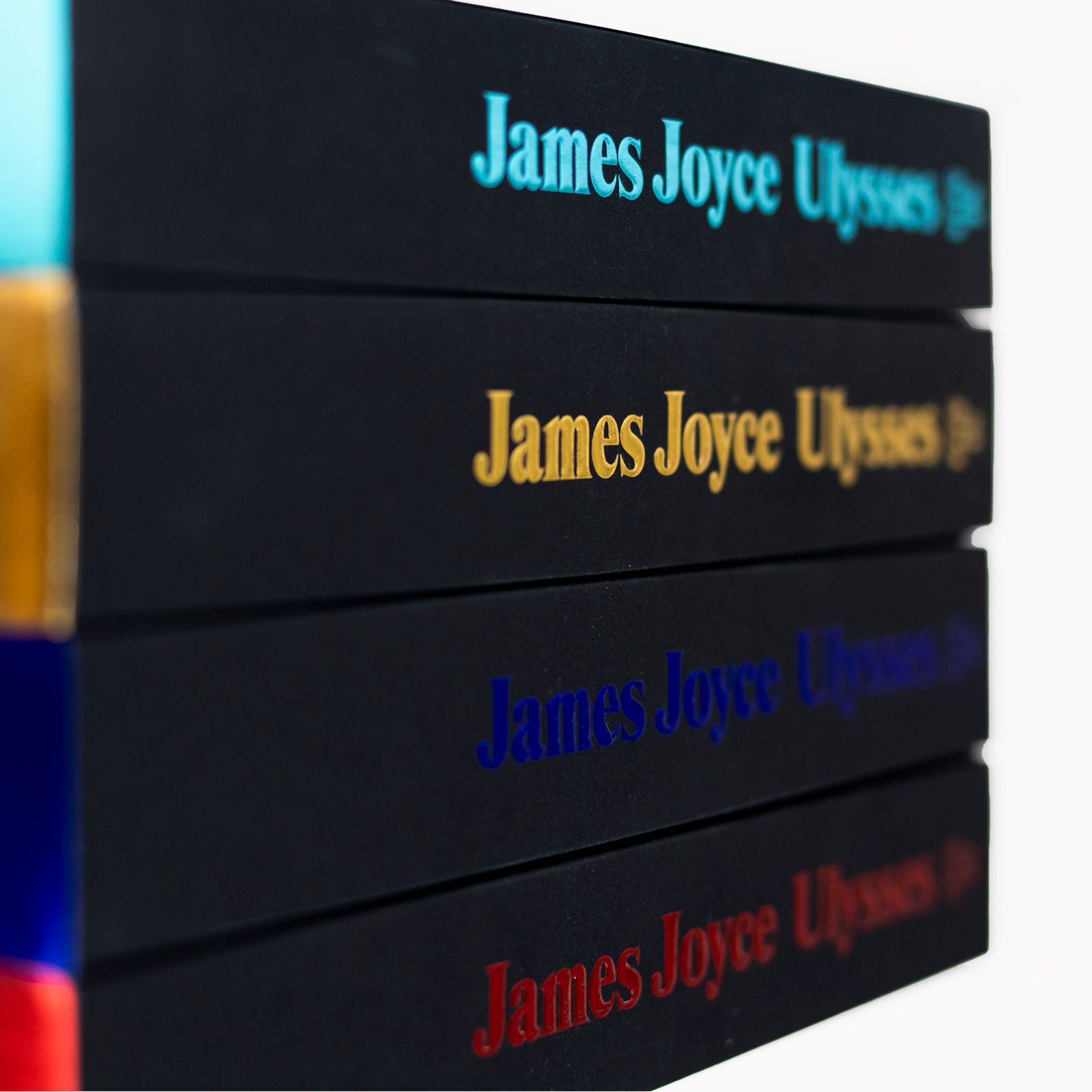 Ein Stapel mit vier Ausgaben des Buches »Ulysses« von James Joyce mit jeweils verschiedenfarbiger Prägung. Die Ausgaben sind von der Seite fotografiert, zu sehen sind die Buchrücken in den Farben Türkis, Blau, Gold und Rot.