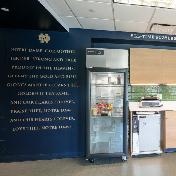 Dimensional lettering spelling Notre Dame Victory March lyrics in gold on navy wall above team kitchenette area.