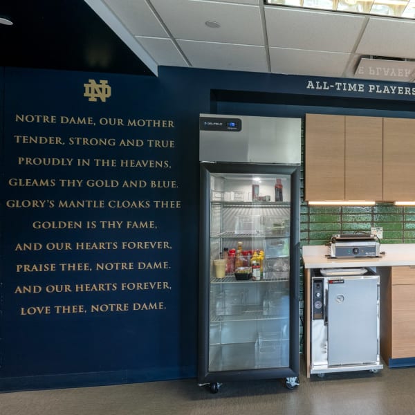 Dimensional lettering in gold spelling Notre Dame Victory March on navy wall beside locker room kitchenette and timeline.