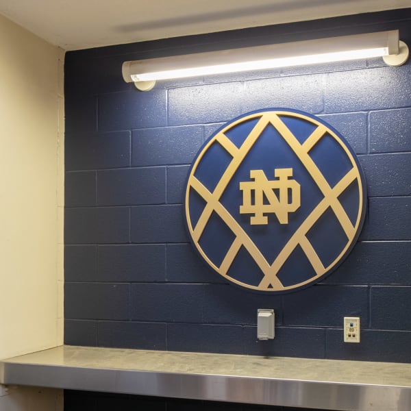 School logo sign featuring dimensional circular lacrosse sticks emblem in gold and navy mounted on painted block wall.