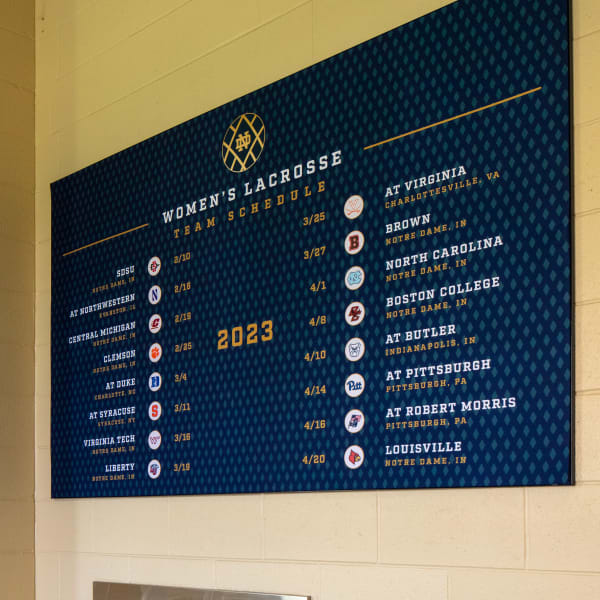Notre Dame stadium sign displaying women's lacrosse 2023 team schedule on textured navy panel above water fountains.
