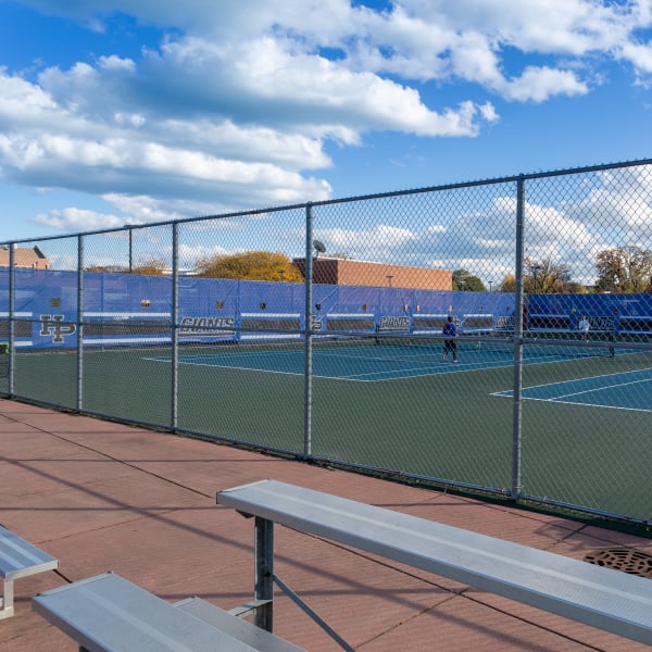Blue windscreen mesh banner installed on chain link fence behind tennis court bench area.