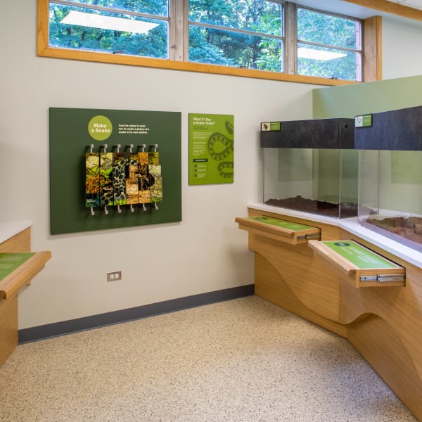 Nature center exhibit room showing glass terrariums, a green spinning photo board, and environmental graphics panels on white walls.