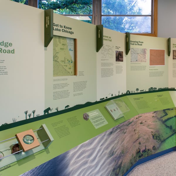 Full view of curved environmental graphics installation with historical aerial photos, timeline panels, and small artifact displays.