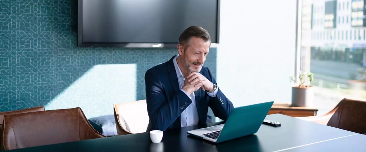 Ein älterer Mann im Anzug sitzt in einem modernen Büro an einem Tisch und schaut konzentriert auf ein aufgeklapptes Laptop. Neben ihm steht eine Kaffeetasse, im Hintergrund ist ein großes Fenster mit Blick auf ein Bürogebäude zu sehen.