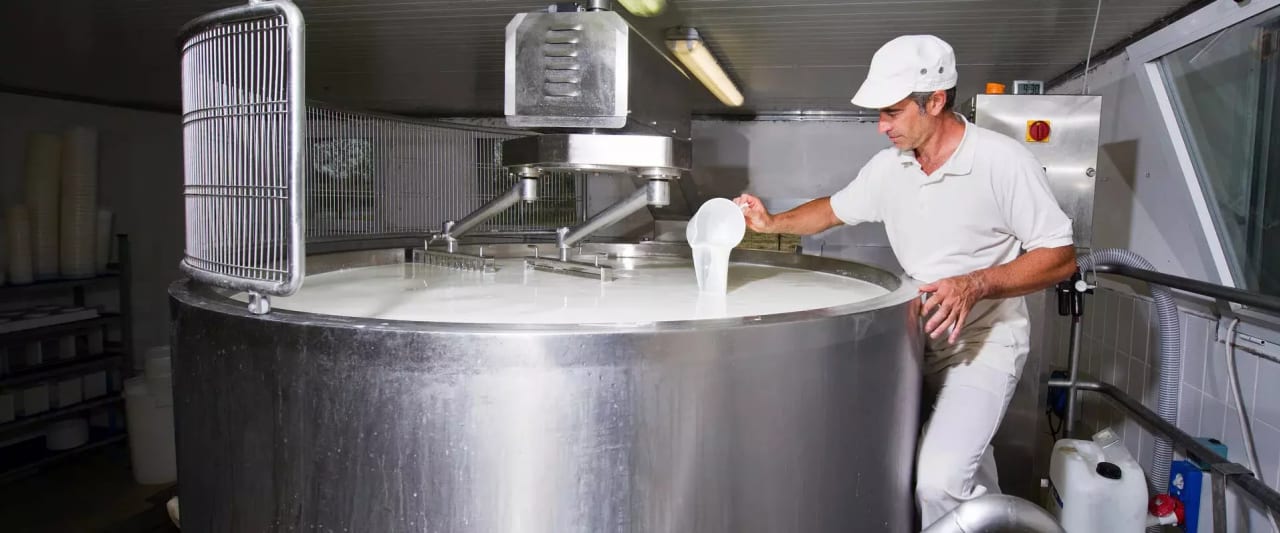 Ein Mann in weißer Arbeitskleidung überprüft mit einem Becher die Flüssigkeit in einem großen Edelstahltank in einer Molkerei oder Käserei.