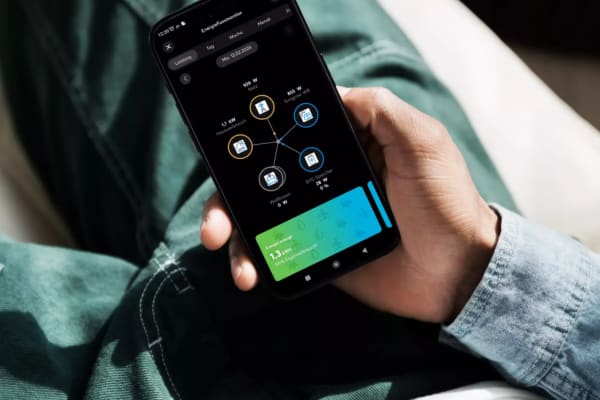 Eine Person h&auml;lt ein Smartphone mit ge&ouml;ffneter Energie-App in der Hand. Auf dem Display ist ein Energieflussmonitor zu sehen, der Stromverbrauch und -quellen visualisiert.