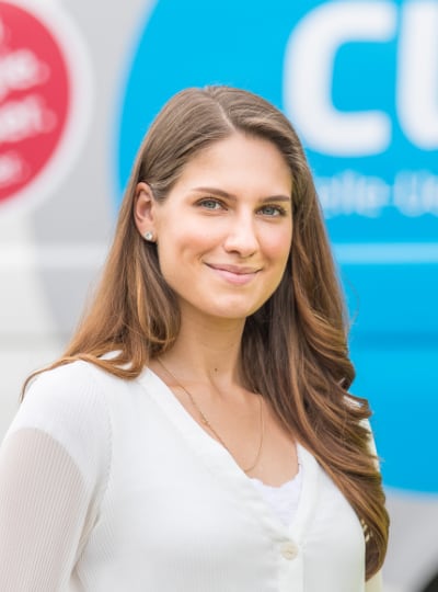 Junge Frau mit langen braunen Haaren und weißer Bluse lächelt in die Kamera, im Hintergrund ein blaues Logo.