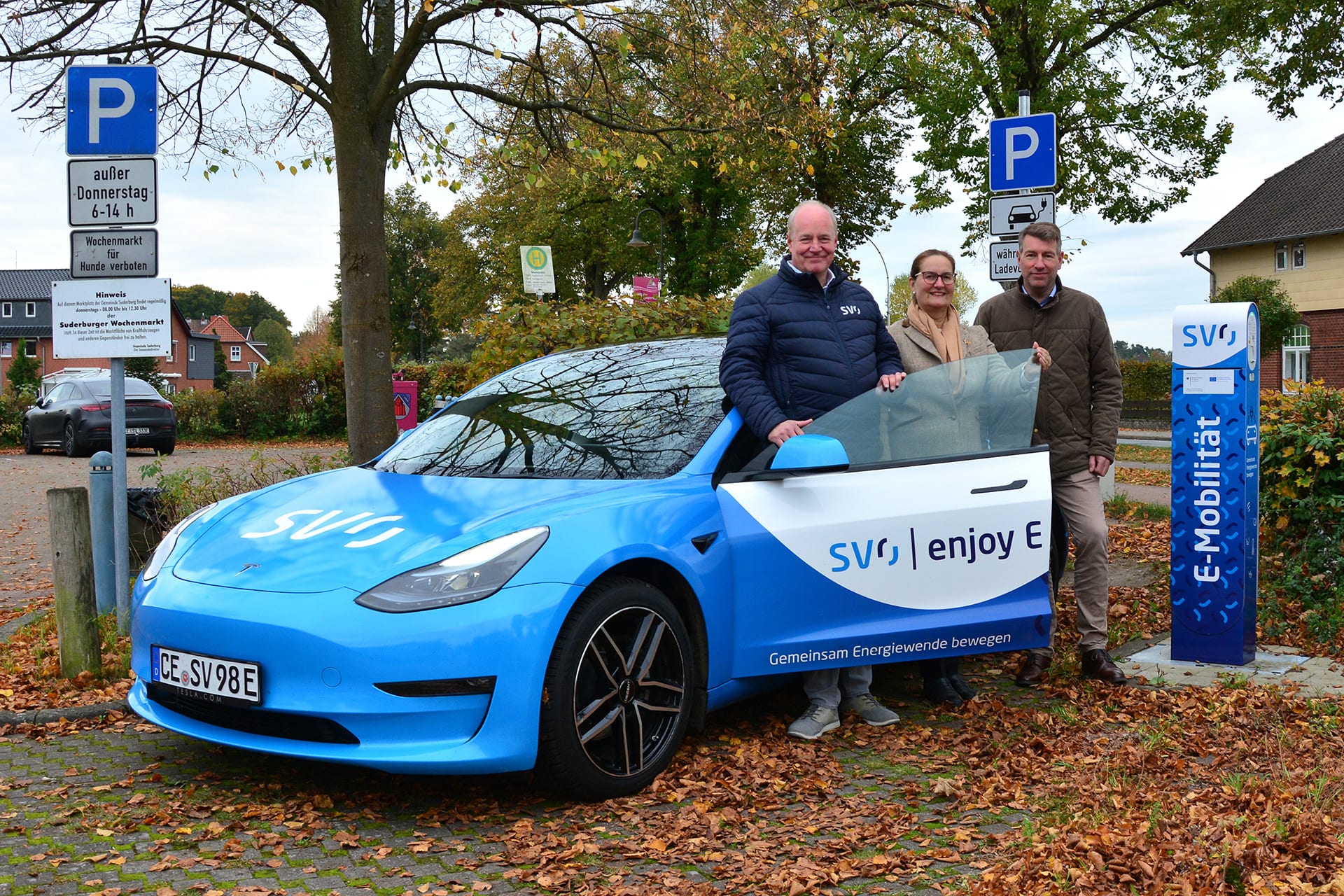 Zwei Männer und eine Frau an einem blauweißen E-Auto neben einer blauweißen Ladesäule.