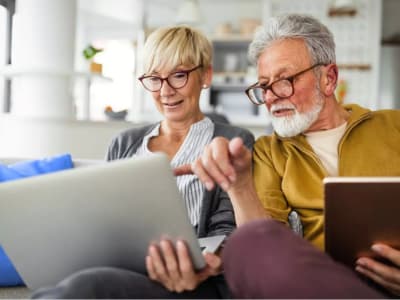 Älteres Paar sitzt gemeinsam auf dem Sofa und schaut interessiert auf einen Laptop.