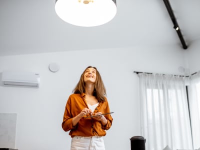Eine Frau steht in einem hellen, modernen Raum und steuert mit einem Tablet die Beleuchtung über ihr. Sie lächelt und blickt zur Lampe an der Decke.