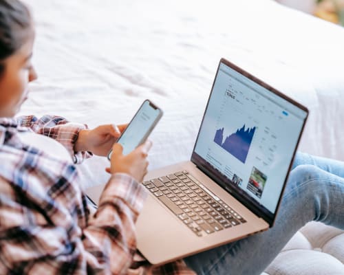 Frau sitzt auf dem Bett und betrachtet Aktienkurse auf einem Laptop, während sie gleichzeitig ein Smartphone in der Hand hält.