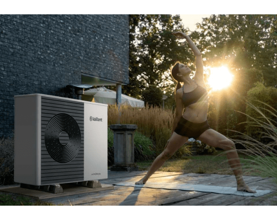 Eine Frau macht bei Sonnenuntergang Yoga im Garten neben einer modernen Wärmepumpe der Marke Vaillant, die neben einem Haus aufgestellt ist.