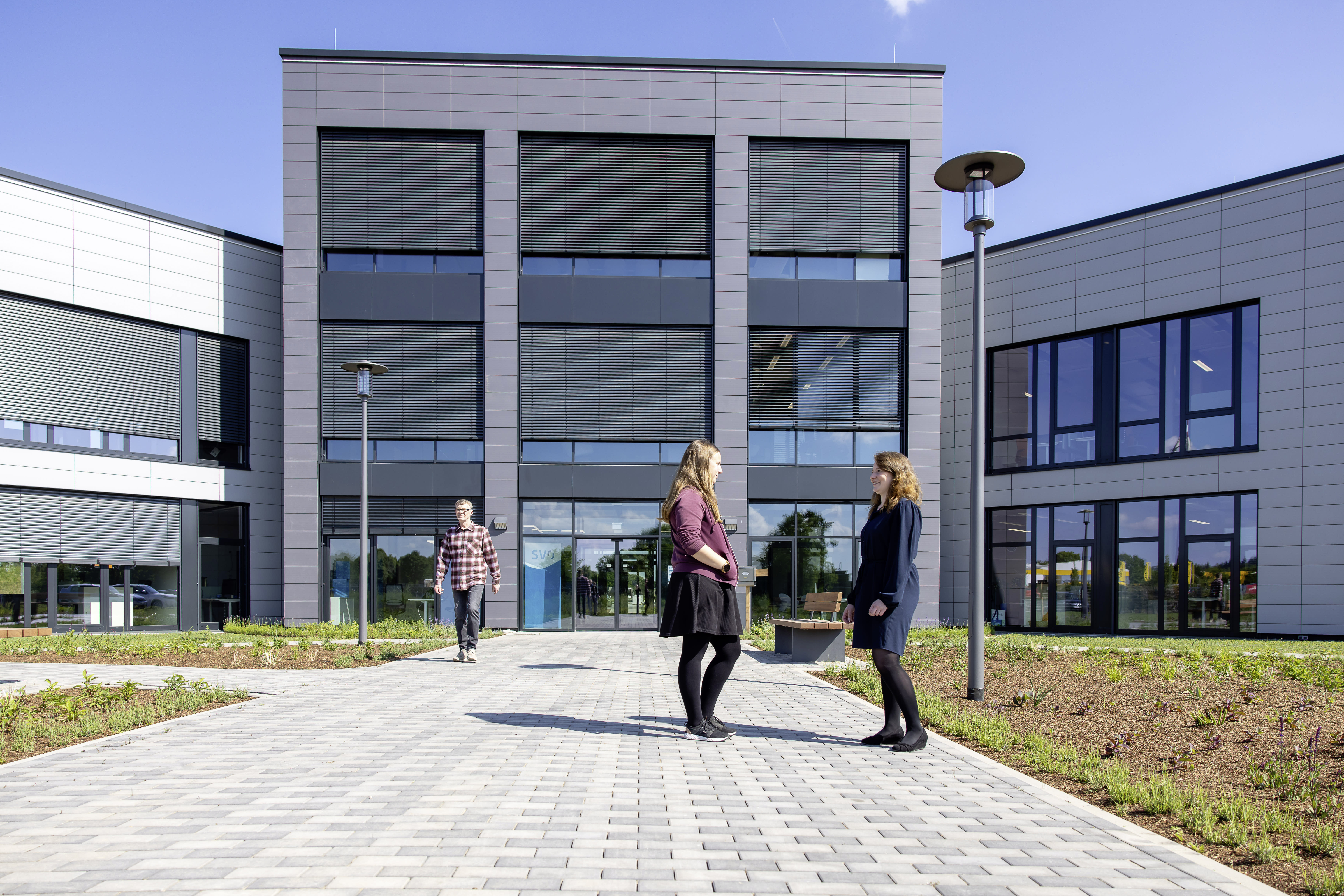 Drei Personen vor einem modernen Bürogebäude: Zwei Frauen unterhalten sich auf dem gepflasterten Weg, ein Mann nähert sich im Hintergrund.