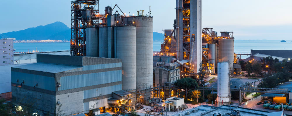 Eine große Industrieanlage bei Abenddämmerung, bestehend aus mehreren Silos, Türmen und Rohrleitungen. Im Hintergrund sind ein Gewässer und Berge zu sehen. Die Anlage ist beleuchtet, was auf laufenden Betrieb hinweist.