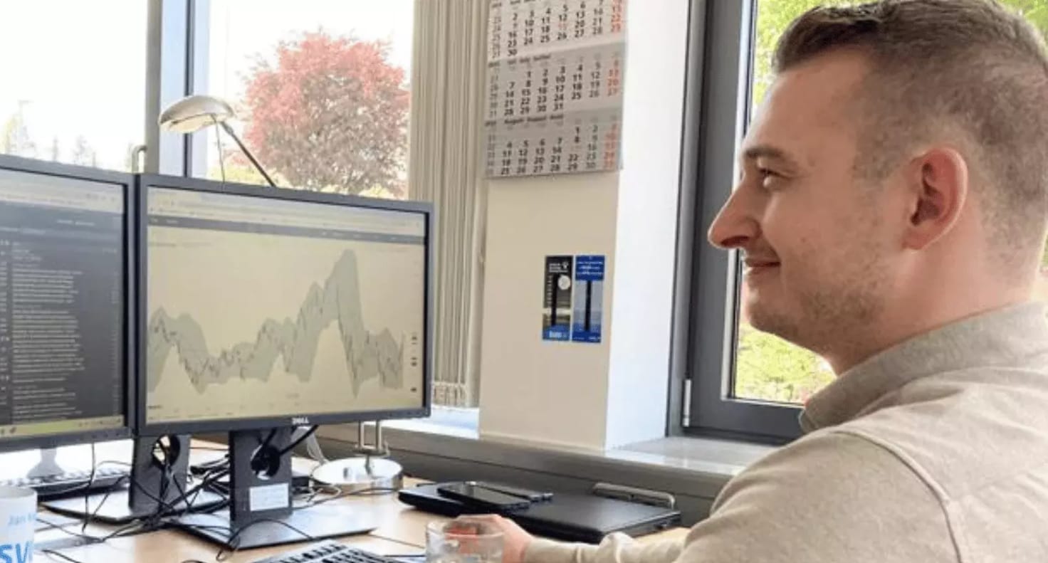 Mitarbeiter sitzt an einem Schreibtisch mit zwei Monitoren, auf denen Marktdaten und Diagramme angezeigt werden.