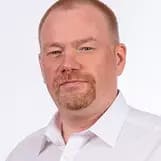 Porträt eines Mannes mit kurzen roten Haaren, Bart und weißem Hemd vor hellem Hintergrund.