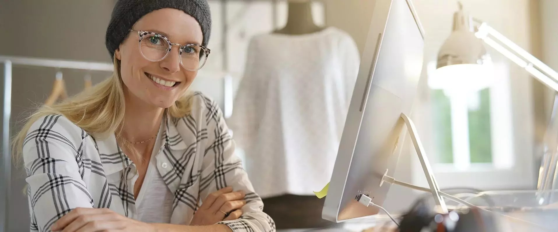 Lächelnde Frau mit Brille und Mütze sitzt an einem Schreibtisch vor einem großen Monitor. Sie trägt ein kariertes Hemd und hat die Arme verschränkt. Im Hintergrund sind ein Schaufenster und eine Schneiderpuppe zu erkennen, was auf ein kreatives oder gestalterisches Arbeitsumfeld hinweist. Die Szene ist hell und freundlich beleuchtet.
