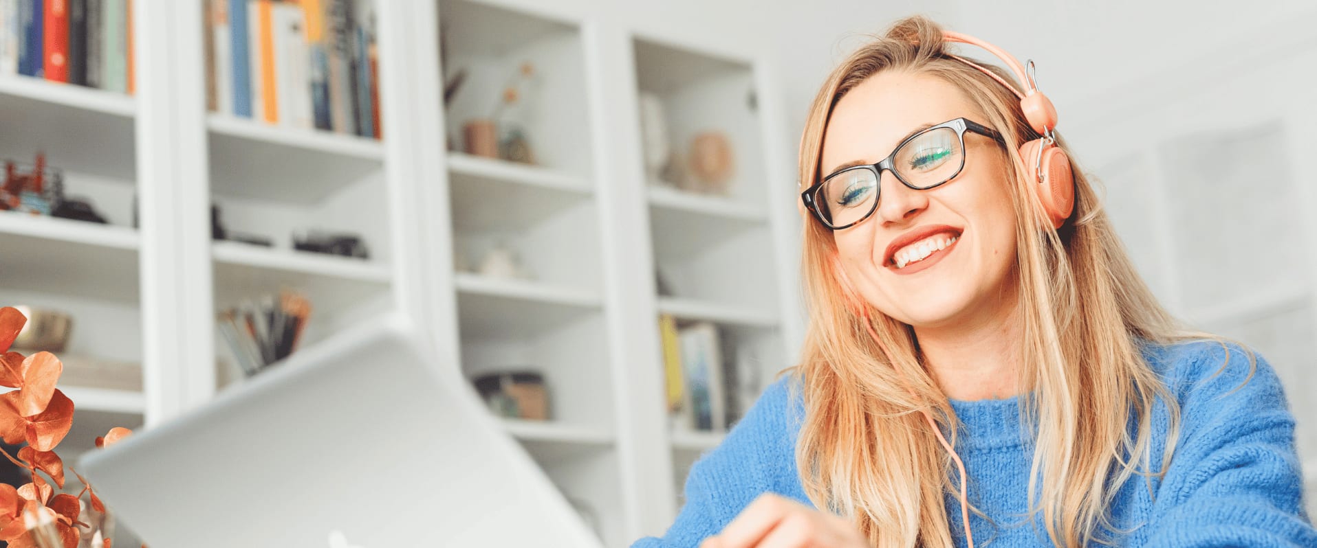 Frau mit Kopfhörern und Brille sitzt lächelnd vor einem Laptop in einem hellen Raum mit Bücherregalen.