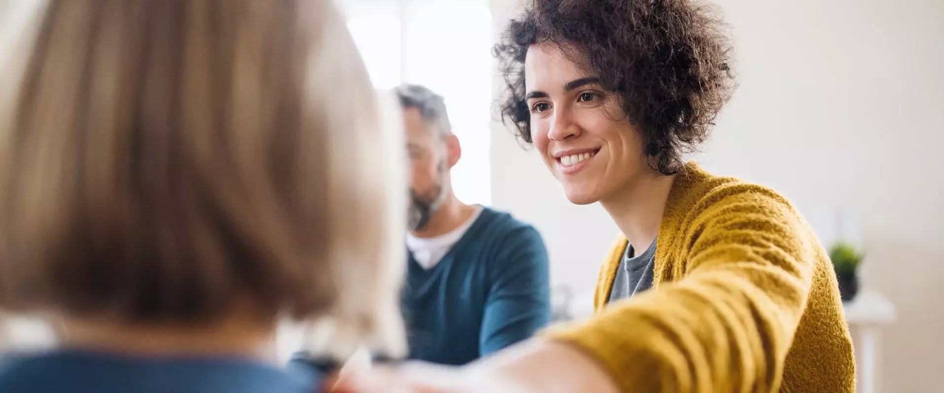 Eine junge Frau mit lockigem Haar und gelber Strickjacke lächelt freundlich und reicht einer anderen Person die Hand, während im Hintergrund ein Mann sitzt.