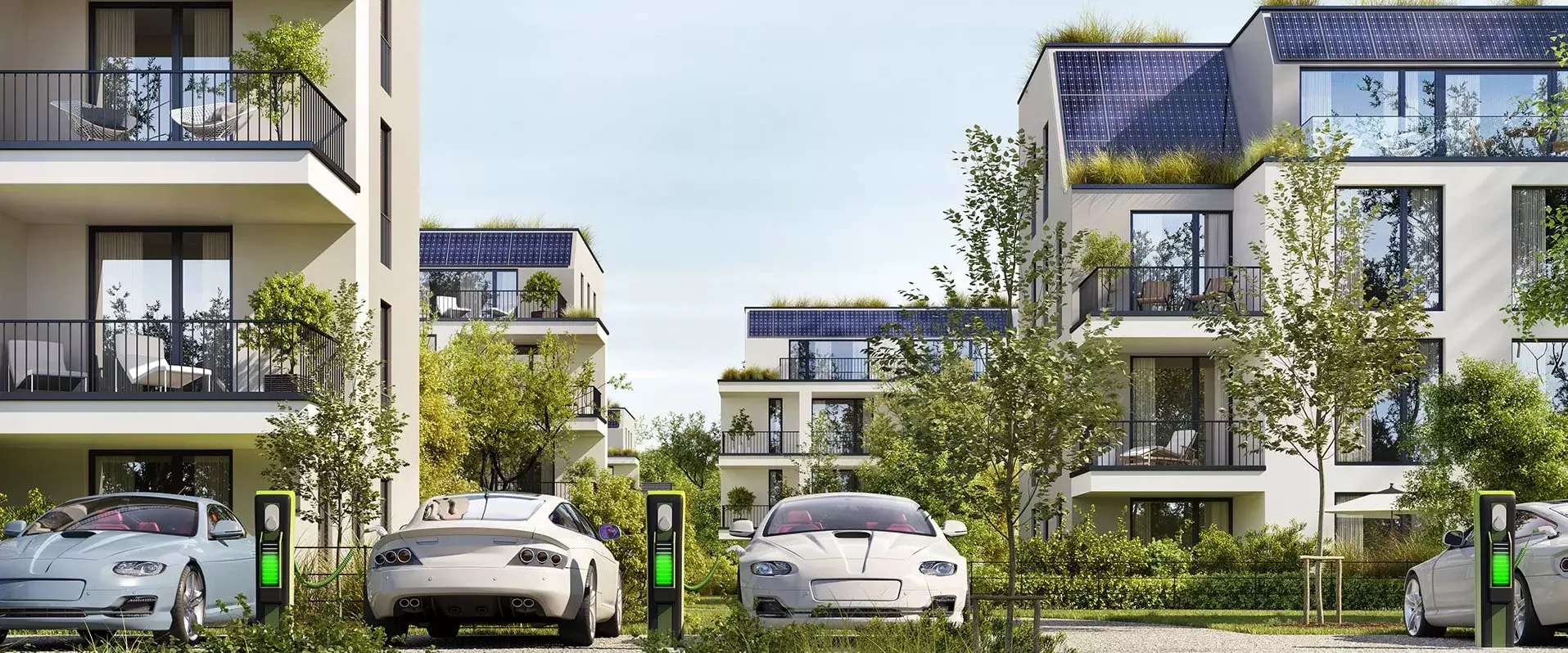 Mehrere moderne Wohngebäude mit begrünten Dächern und Solarpanels. Davor stehen Elektroautos an Ladestationen, umgeben von viel Grün und Bäumen.