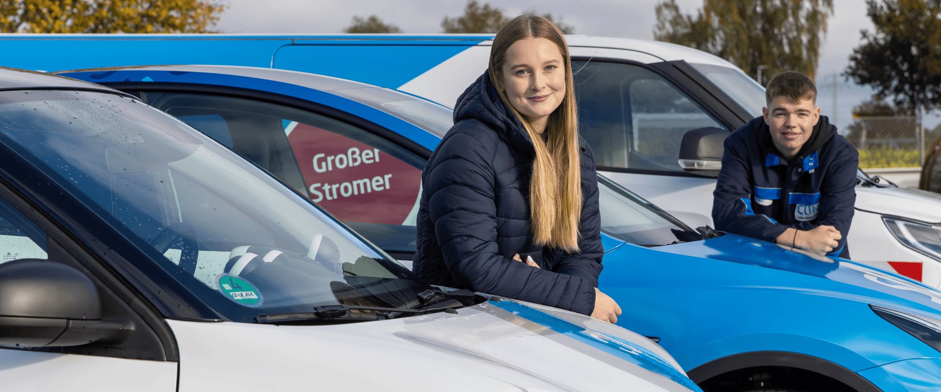Zwei junge Menschen lehnen lächelnd an Elektrofahrzeugen mit SVO-Logo, die nebeneinander auf einem Hof stehen. Im Hintergrund ist ein blau-weißer Transporter mit der Aufschrift „Großer Stromer“ sowie eine große leere Kabeltrommel zu sehen.