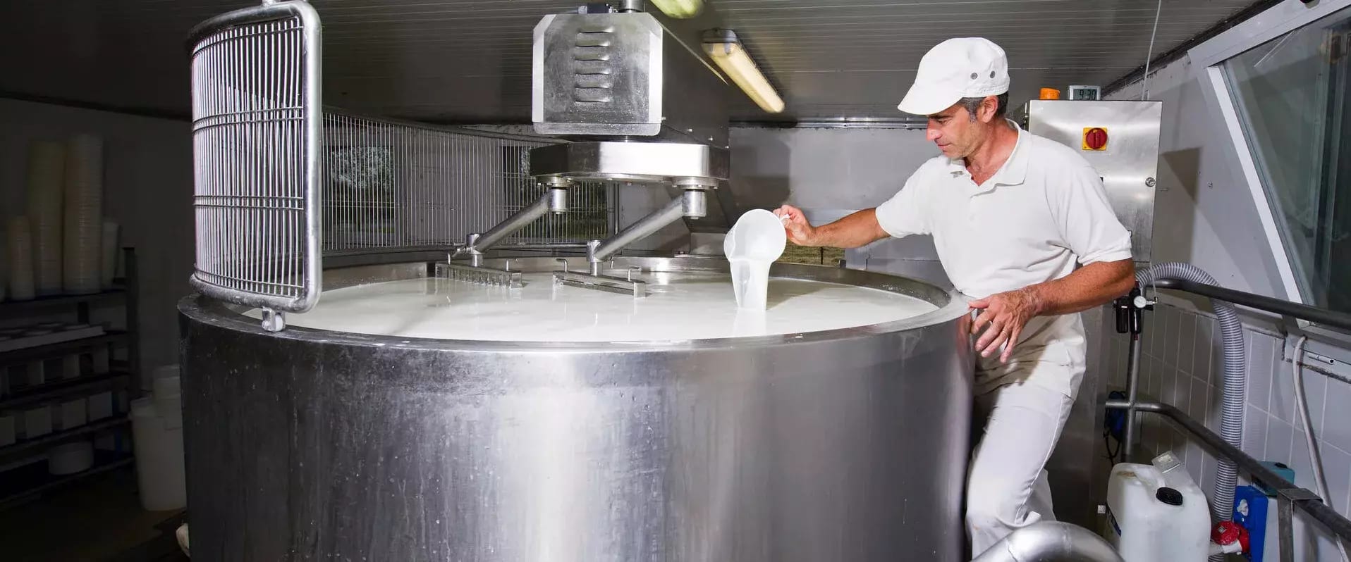 Ein Mann in weißer Arbeitskleidung überprüft mit einem Becher die Flüssigkeit in einem großen Edelstahltank in einer Molkerei oder Käserei.