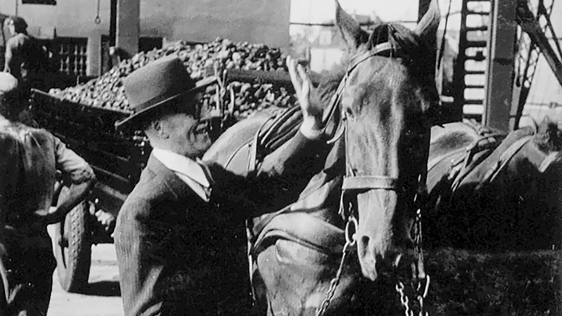 Mann mit Hut streichelt ein Pferd vor einem beladenen Wagen in historischer Aufnahme.