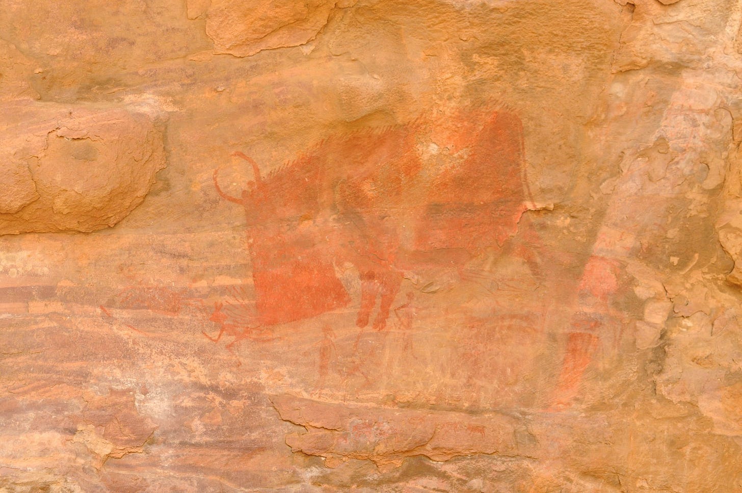 Prehistoric rock art in deep red showing a massive stylized animal, identified as a boar or bison, charging toward small human figures on a tan rock wall.