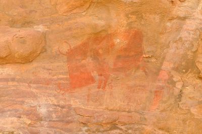 Prehistoric rock art in deep red showing a massive stylized animal, identified as a boar or bison, charging toward small human figures on a tan rock wall.
