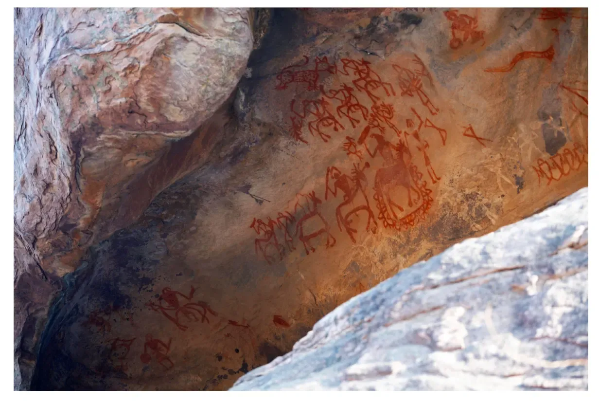 A wide photograph showing red ochre rock paintings under a natural stone overhang at the Bhimbetka Rock Shelters, depicting stick figures, horseback riders, hunters with bows, and dancing figures drawn on sandstone walls.