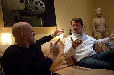 Harvard psychologists Daniel Gilbert (left) and Matthew Killingsworth (right) sitting on a living room sofa, engaged in an animated, expressive discussion. Gilbert, bald with glasses and a black shirt, points open-handed while Killingsworth, wearing a white button-down shirt and jeans, gestures back while explaining their research. A large black-and-white print of a vintage film projector hangs on the wall behind them alongside a warm lamp and stone sculpture.