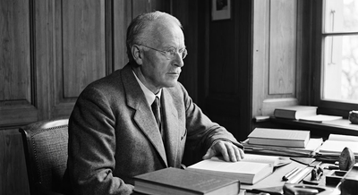 A black-and-white portrait of Carl Gustav Jung in a suit and round spectacles, seated at a wooden desk in his study while resting his hand on an open book.