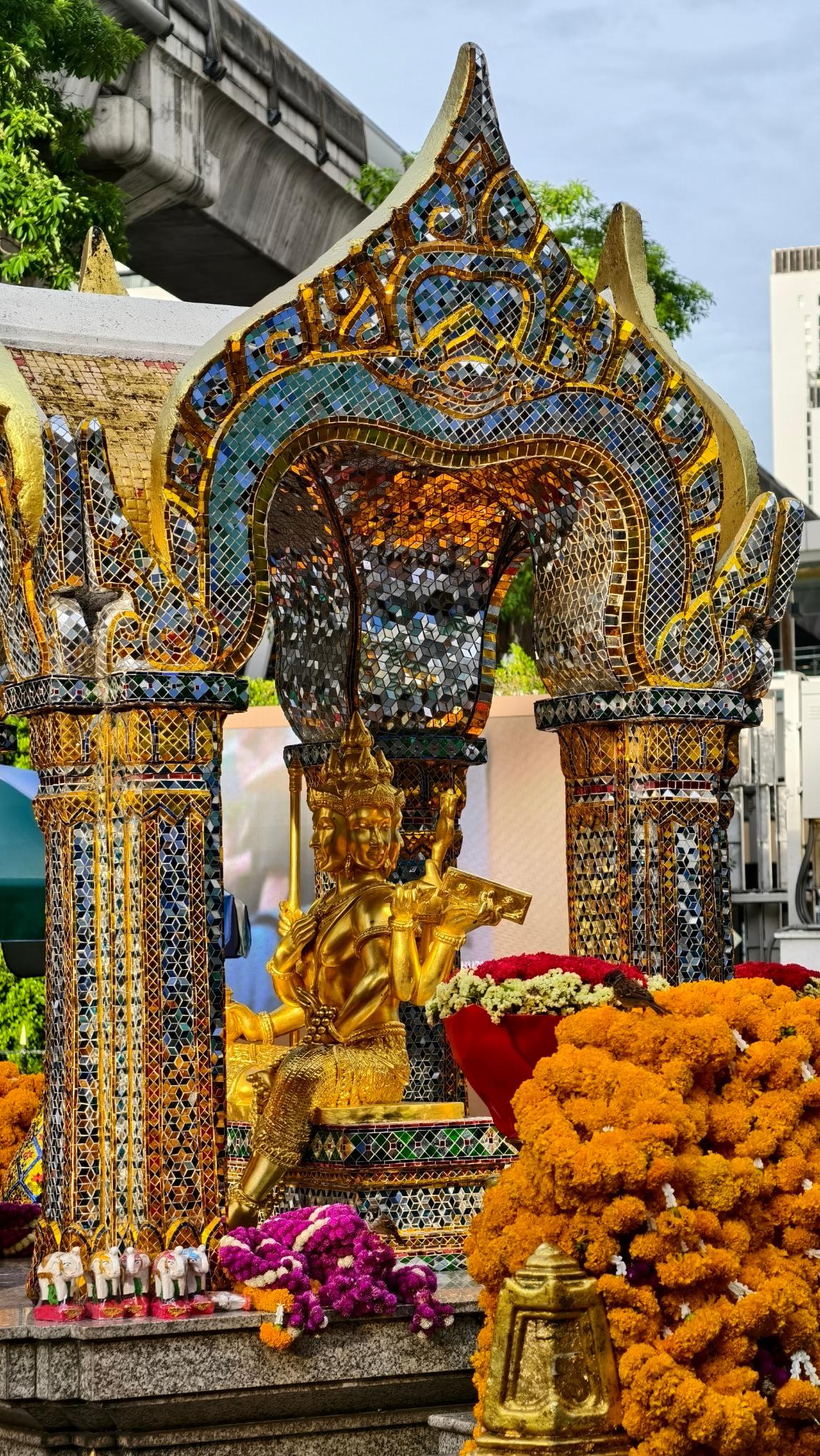 ศาลาพระพรหมเอราวัณยามเย็น มีพวงมาลัยดอกดาวเรืองและช้างไม้แกะสลักวางถวาย