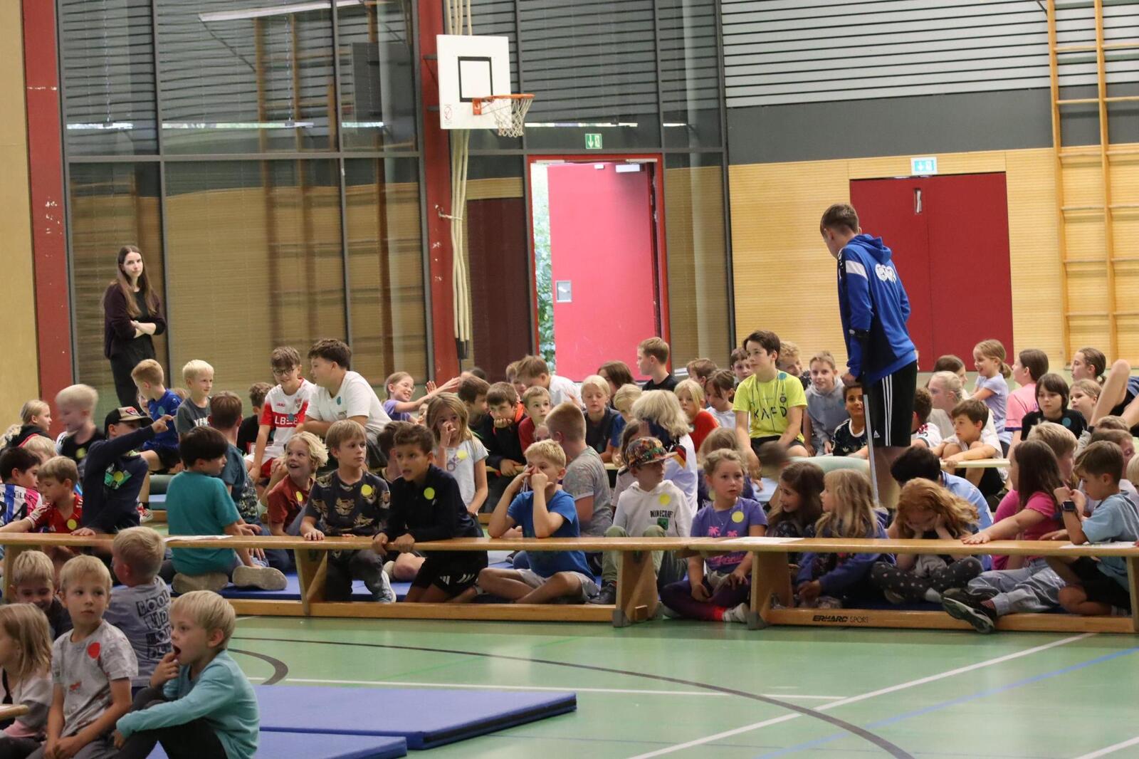Die Kinderschar sitzt auf Bänken und Matten in der Halle