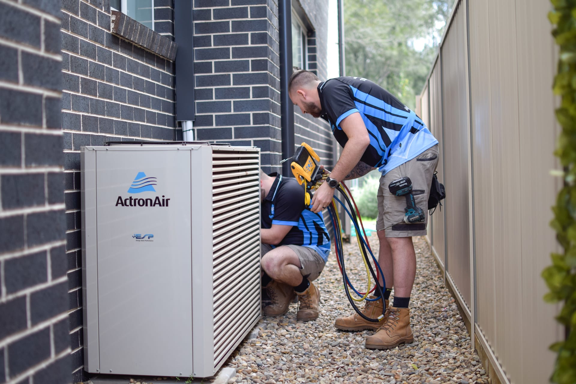 Two professional air conditioning technicians servicing a residential outdoor condenser with manifold gauges beside a modern brick home