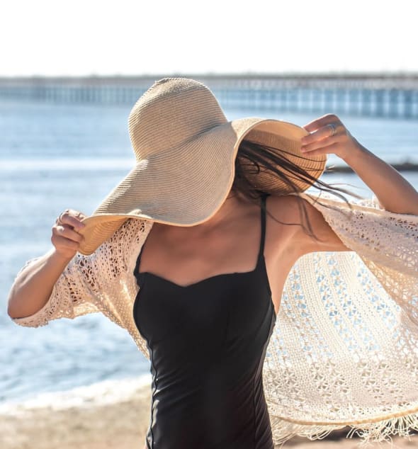 A woman wearing a black swimsuit, a wide-brimmed straw hat, and a light crocheted cover-up, with the ocean and a distant pier in the background