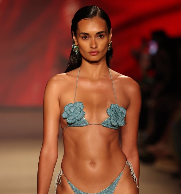 Model walking the runway in a shimmering teal bikini with floral appliqués, styled with bold earrings against a vibrant background.