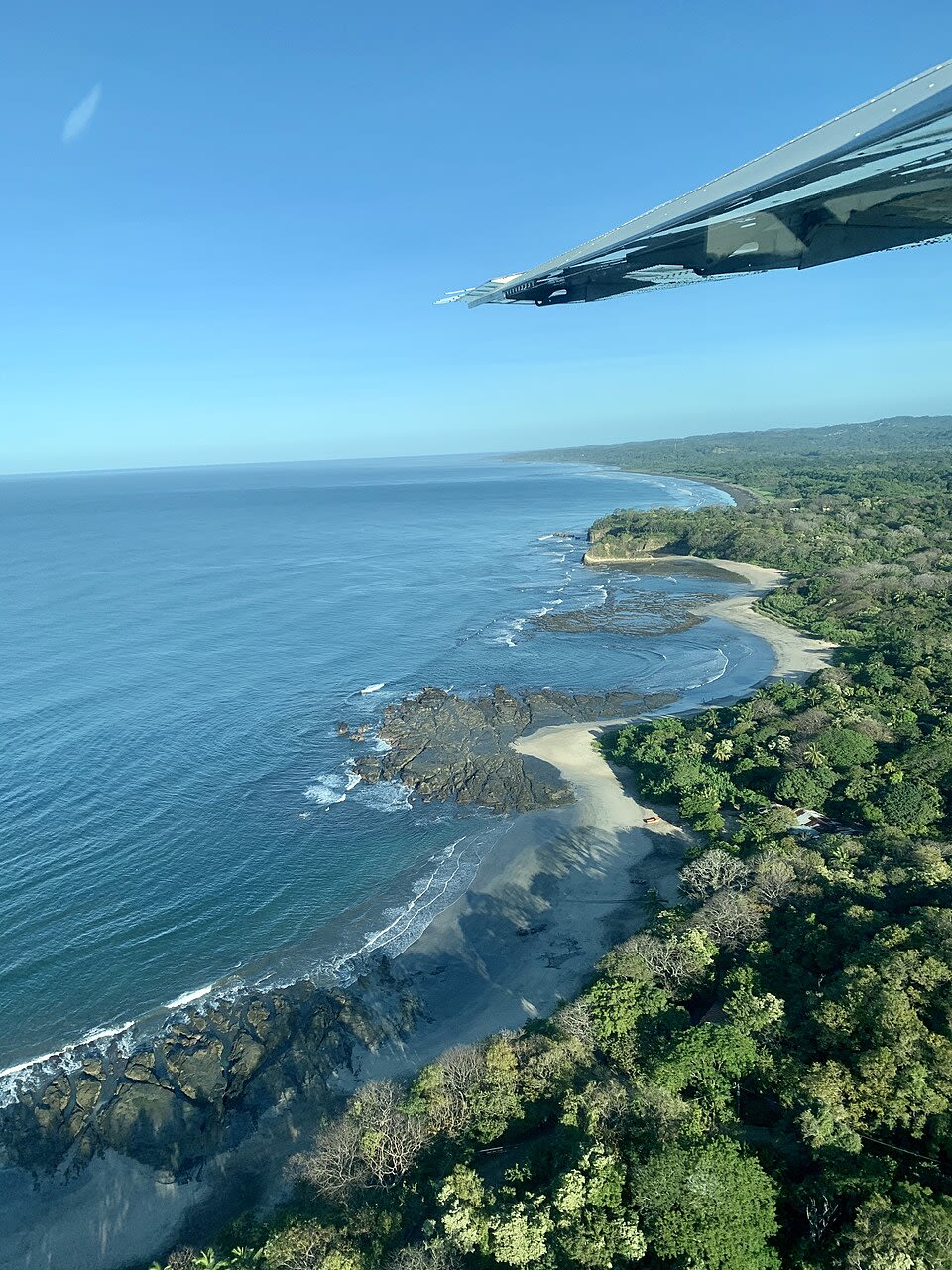 Aerial view of a serene coastline featuring rocky outcrops and soft sandy beaches, surrounded by lush green vegetation under a clear blue sky.