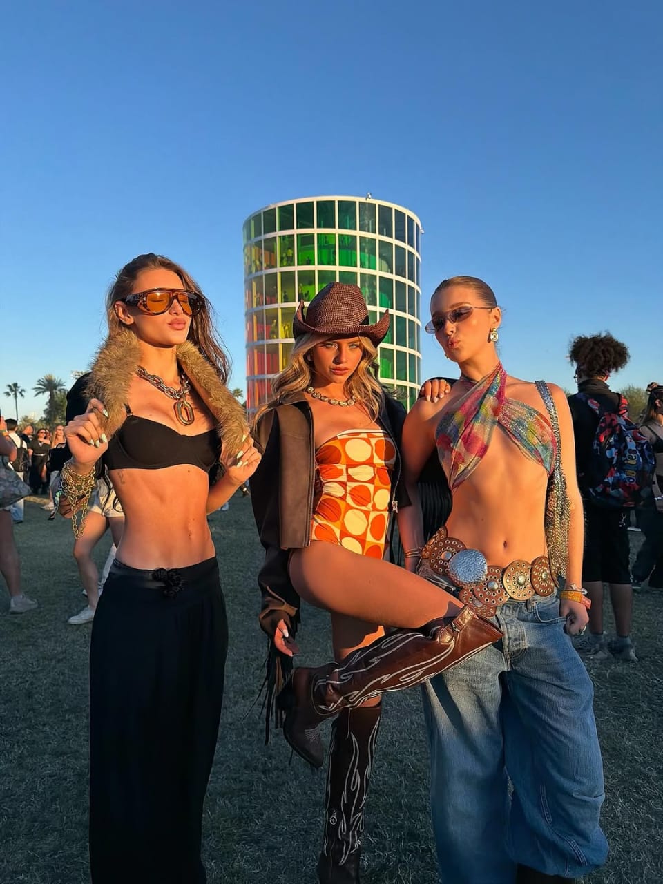 Three stylish women pose together at Coachella 2k26, showcasing bold swimwear fashion, including vibrant prints and accessories against a colorful backdrop.