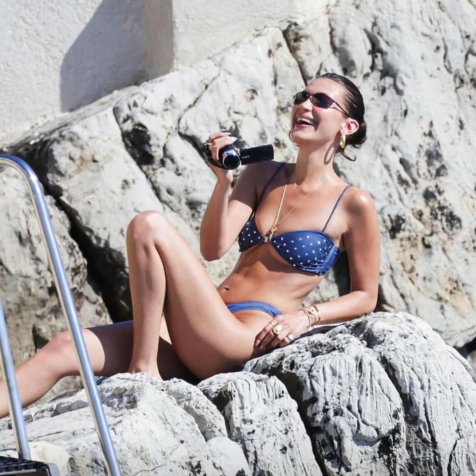 A woman in a blue polka dot bikini relaxes on sunlit rocks, holding a camera and smiling, accessorized with gold jewelry and sunglasses.