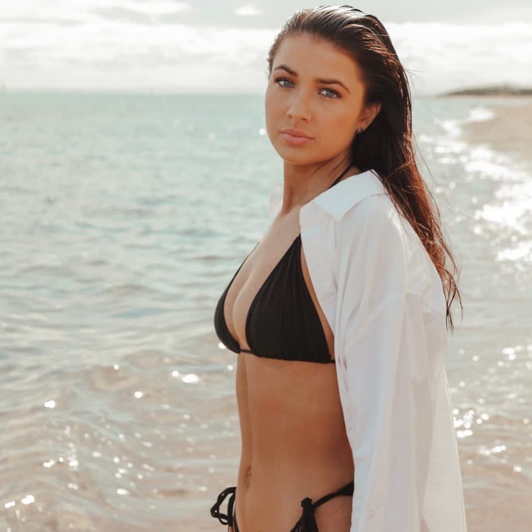 Photo of Janaya Reimers on the beach wearing a black bikini and a white shits