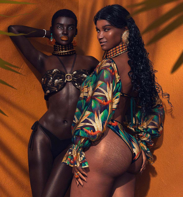 Two women in vibrant swimwear pose against an orange wall; one in a metallic bronze bikini with layered choker, the other in a colorful tropical cover-up and matching bikini, both with bold earrings and glossy, sun-kissed skin.