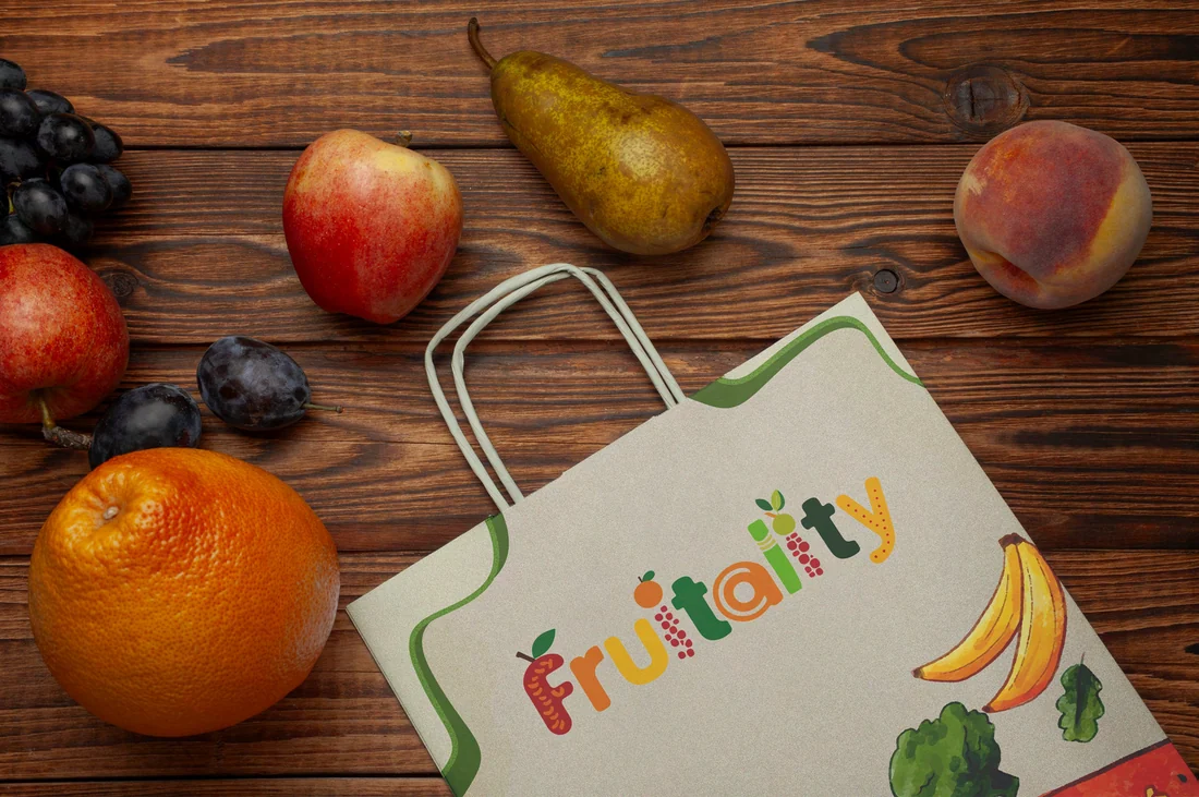 A paper Fruitality carry bag with the wordmark and a green leaf pattern, on a wooden table with apples, pears, an orange and bananas
