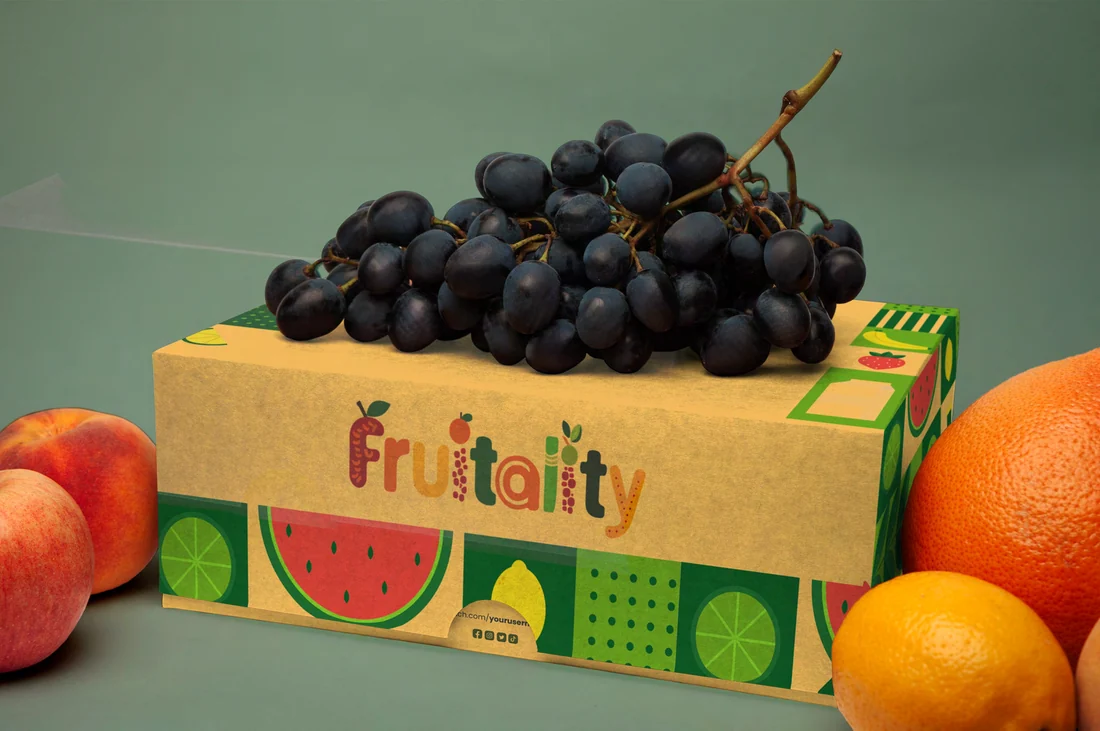 A kraft Fruitality fruit crate with the wordmark and a band of watermelon and lemon slices, grapes piled on top, beside peaches and an orange