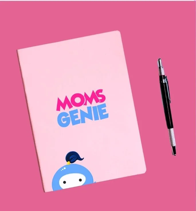 A pink MomsGenie notebook with the wordmark on the cover and the top of Wishie’s head peeking up from the bottom edge