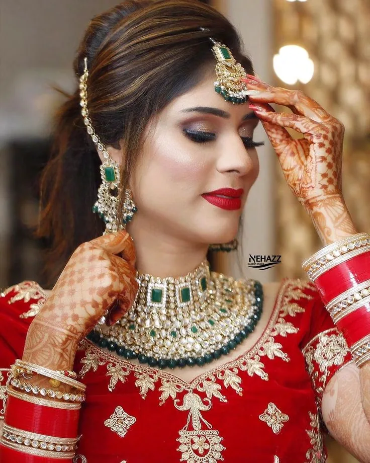 Nehaz Bridal Makeup content: a bride in red and gold with emerald jewellery, eyes lowered, one henna-covered hand raised to her maang tikka
