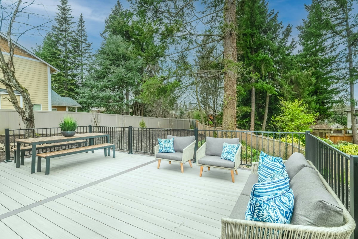 Gray composite deck with an outdoor sofa, chairs, dining table and black railing among evergreen trees